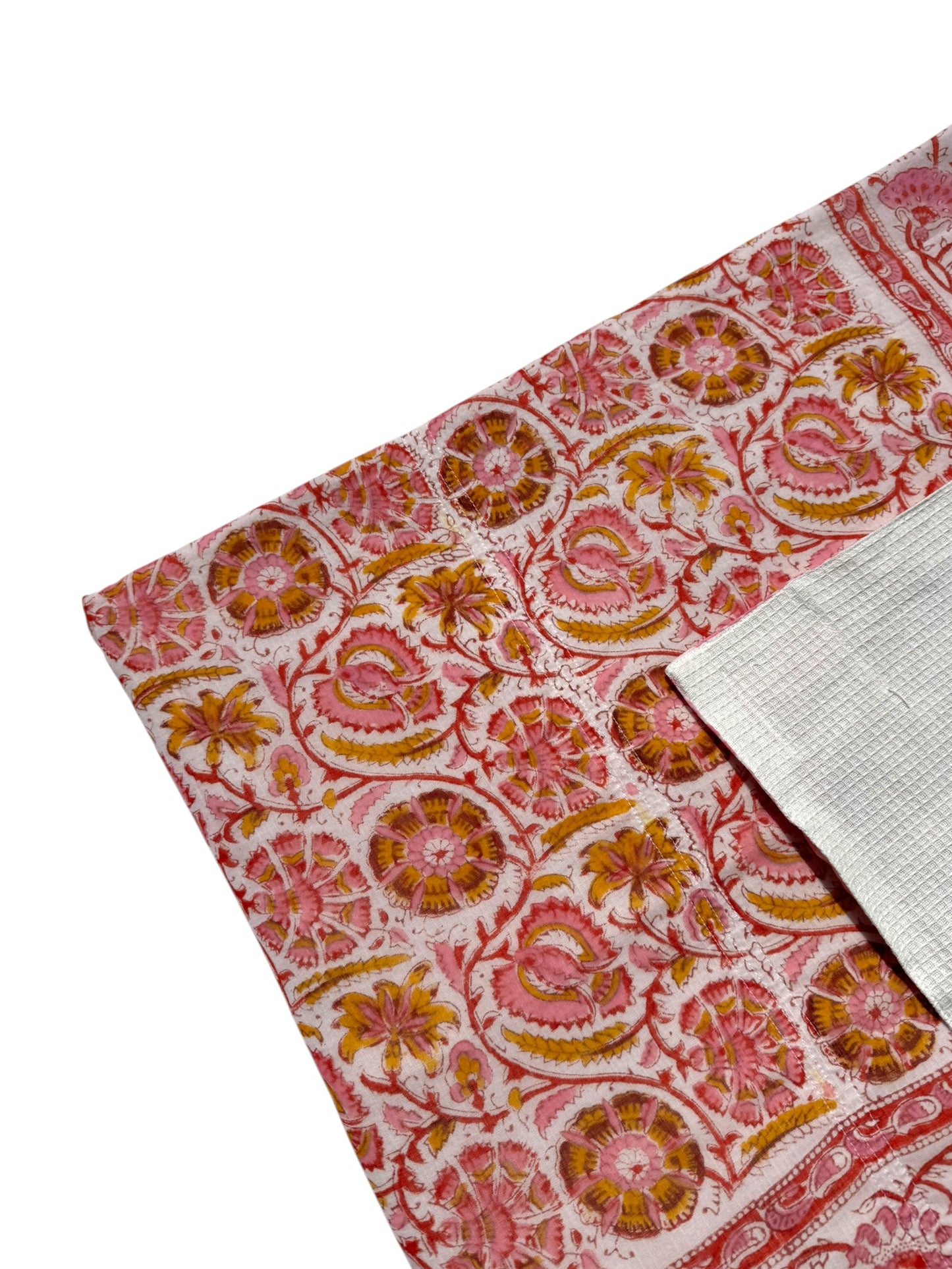 A close-up of a folded textile with an intricate floral and paisley block-print pattern in pink, orange, and yellow on a white base, with a folded white fabric piece showing its woven texture.
