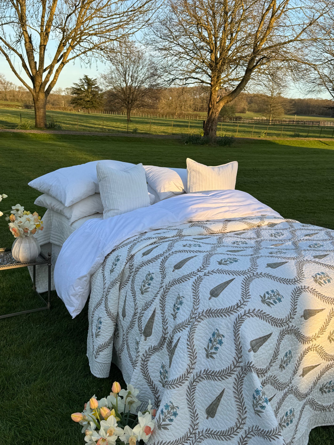 Outdoor bed setup with white bedding and patterned quilt in a grassy field.