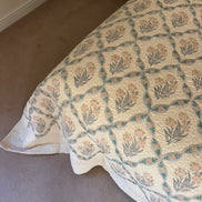 A quilted bedspread with a repeating floral and geometric pattern in muted teal, peach, and gray on an off-white background, styled on a bed in a bedroom with beige carpet.