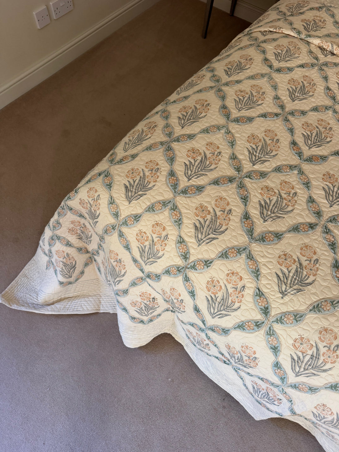 A quilted bedspread with a repeating floral and geometric pattern in muted teal, peach, and gray on an off-white background, styled on a bed in a bedroom with beige carpet.