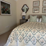 A bedroom scene showing a bed made up with a cream quilt featuring a blue and beige botanical diamond pattern, styled with matching patterned pillows. The room has beige walls, a gray nightstand, a blue ceramic table lamp, and framed wall art.