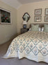 A bedroom scene showing a bed made up with a cream quilt featuring a blue and beige botanical diamond pattern, styled with matching patterned pillows. The room has beige walls, a gray nightstand, a blue ceramic table lamp, and framed wall art.