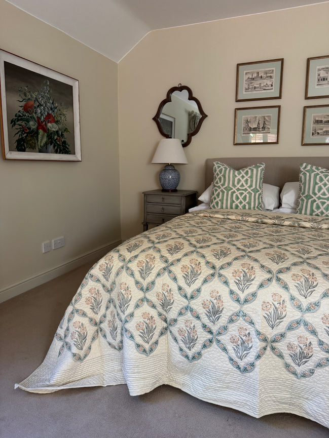 A bedroom scene showing a bed made up with a cream quilt featuring a blue and beige botanical diamond pattern, styled with matching patterned pillows. The room has beige walls, a gray nightstand, a blue ceramic table lamp, and framed wall art.