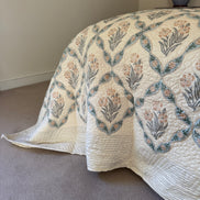A cream quilted bedspread with a botanical diamond pattern in muted blue-green and peach, draped over a bed in a bedroom setting with carpet and a bedside lamp visible.