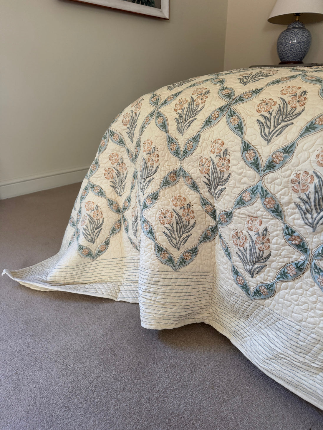 A cream quilted bedspread with a botanical diamond pattern in muted blue-green and peach, draped over a bed in a bedroom setting with carpet and a bedside lamp visible.