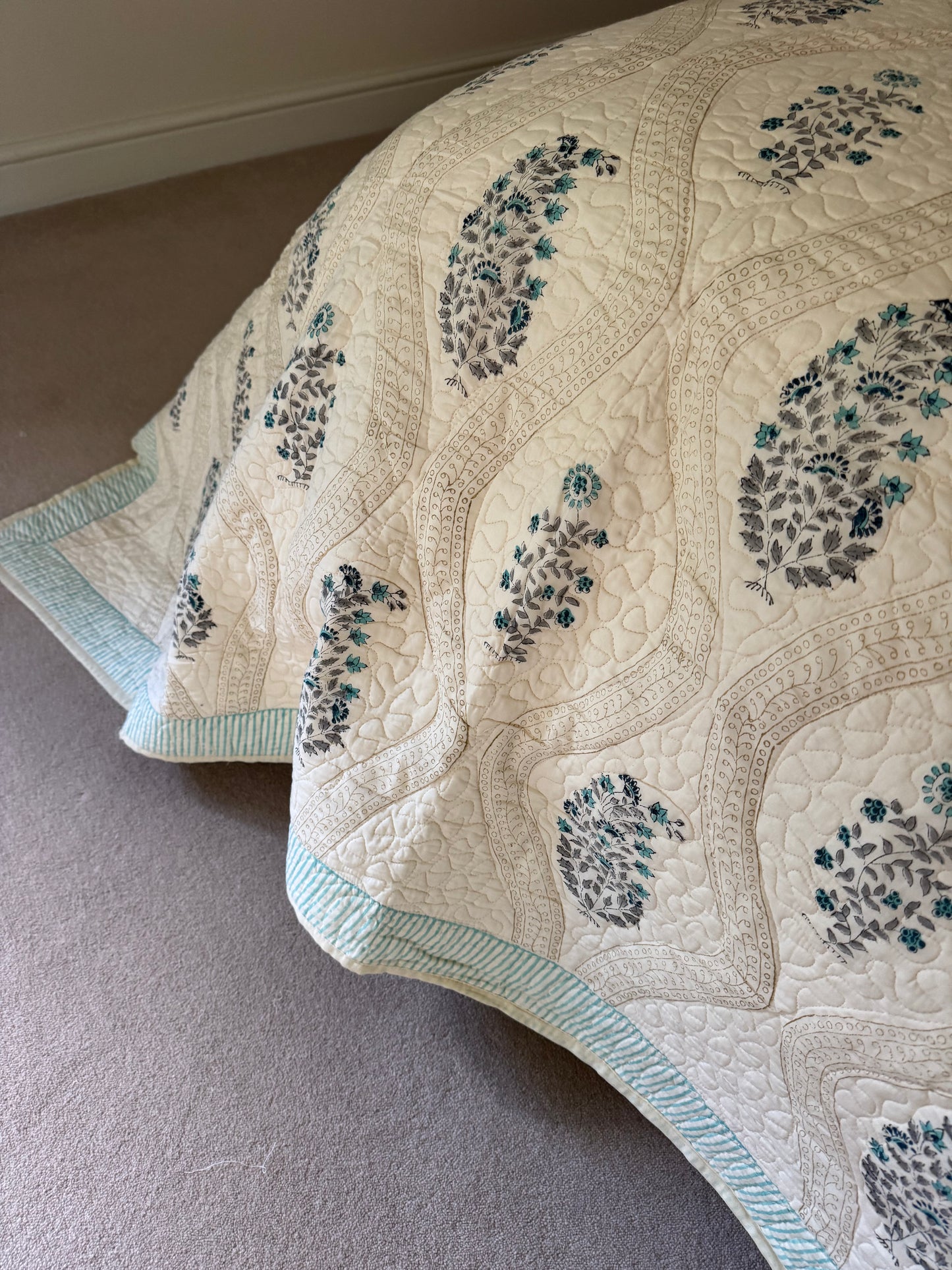 A close-up of a quilted blanket or comforter with a cream base, featuring embroidered floral motifs in teal and gray, and a light blue-green striped border, draped over a bed on a carpeted floor.