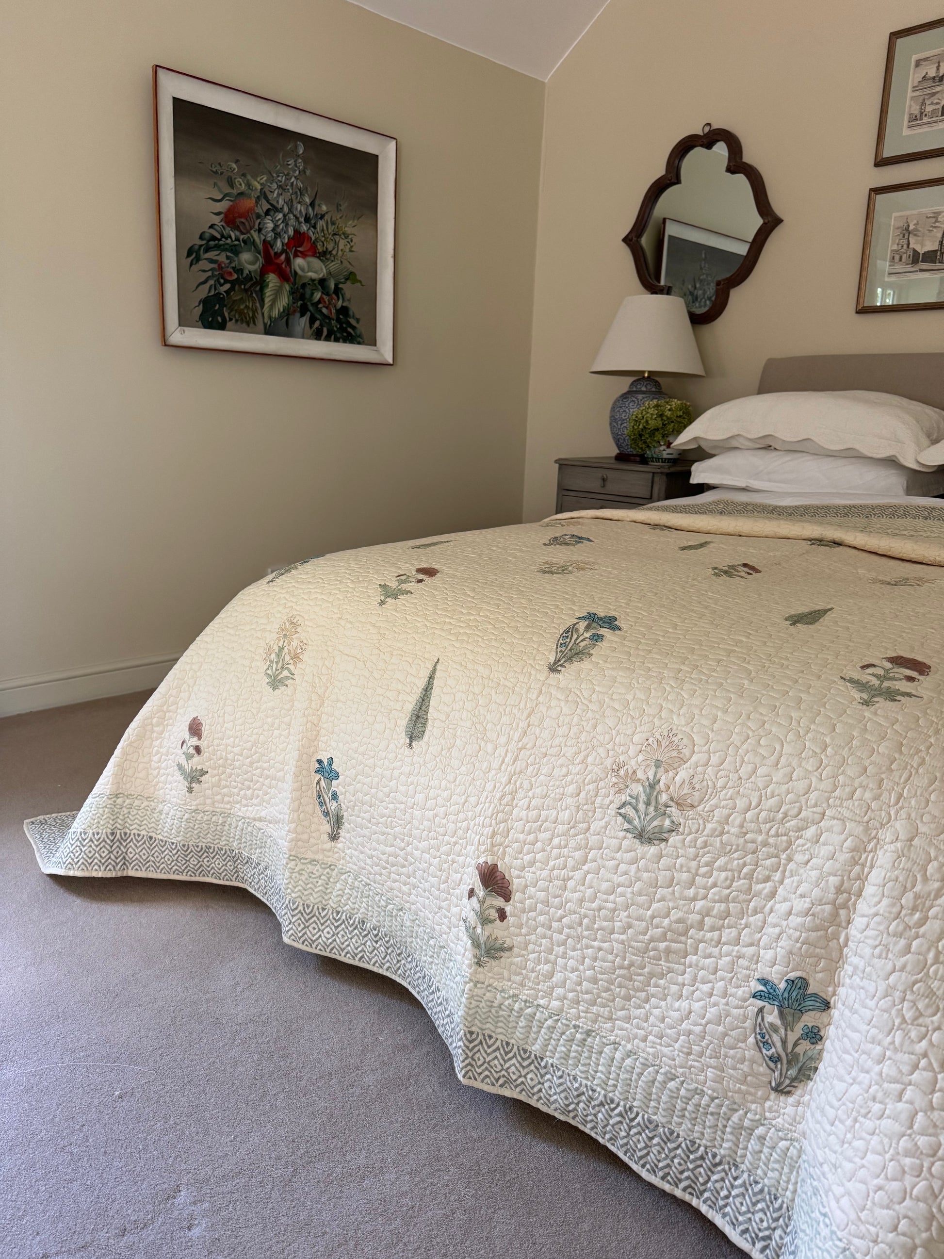 A cream quilted bedspread with a delicate botanical pattern in muted blue, green, and brown tones, styled on a bed in a bedroom setting.