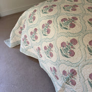 A quilted bedspread with a repeating floral pattern in dusty pink and green on an off-white background, draped over a bed in a bedroom setting with carpeted floor and a beige wall.