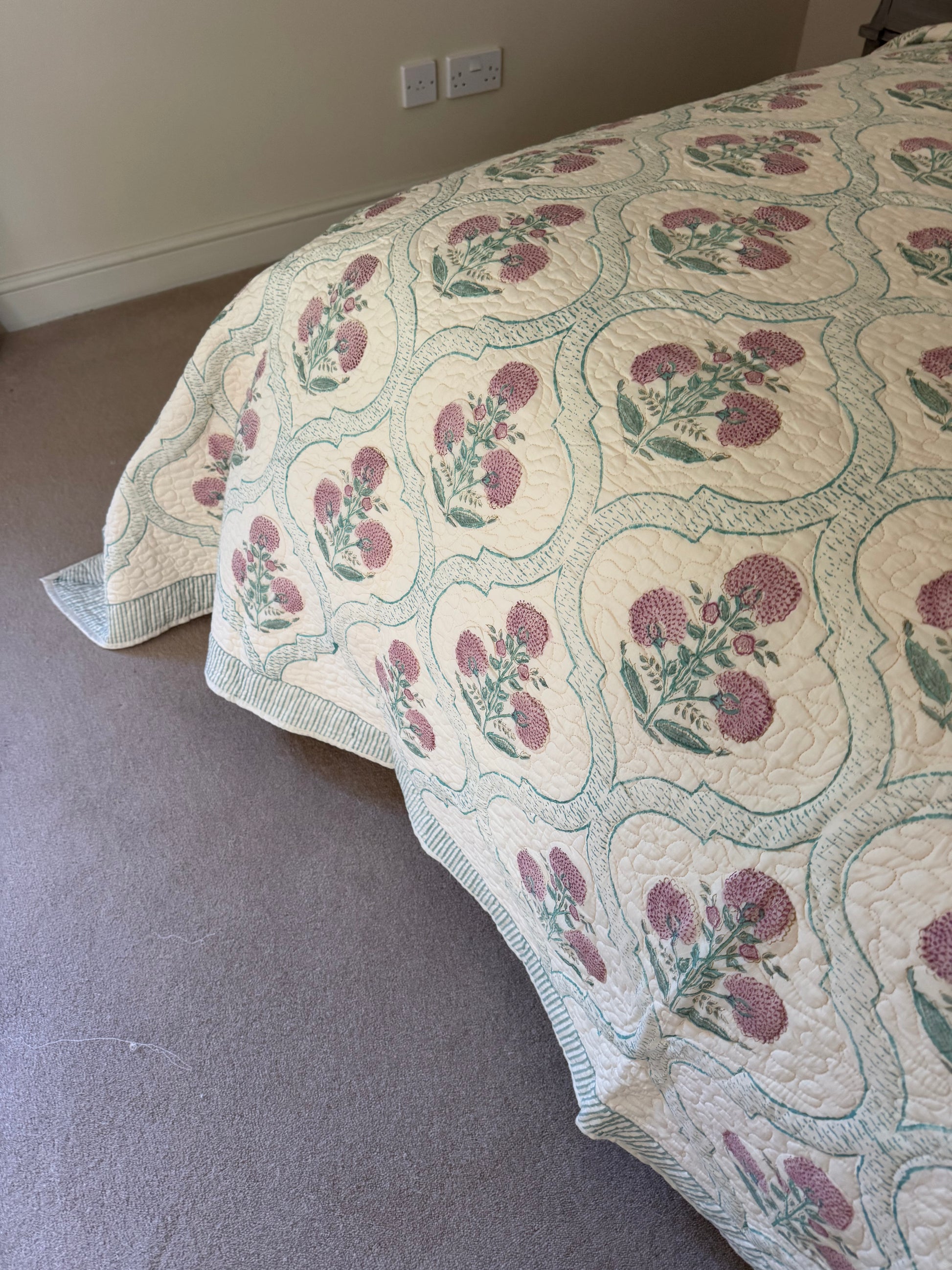 A quilted bedspread with a repeating floral pattern in dusty pink and green on an off-white background, draped over a bed in a bedroom setting with carpeted floor and a beige wall.