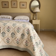 A quilted cream-colored bedspread with a green and pink floral pattern, styled on a bed in a warm, neutral-toned bedroom.