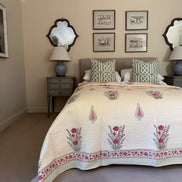A styled bedroom scene featuring a cream quilted bedspread with pink floral motifs and green geometric accent patterns, styled on a bed with matching pillows in a neutral-toned room.