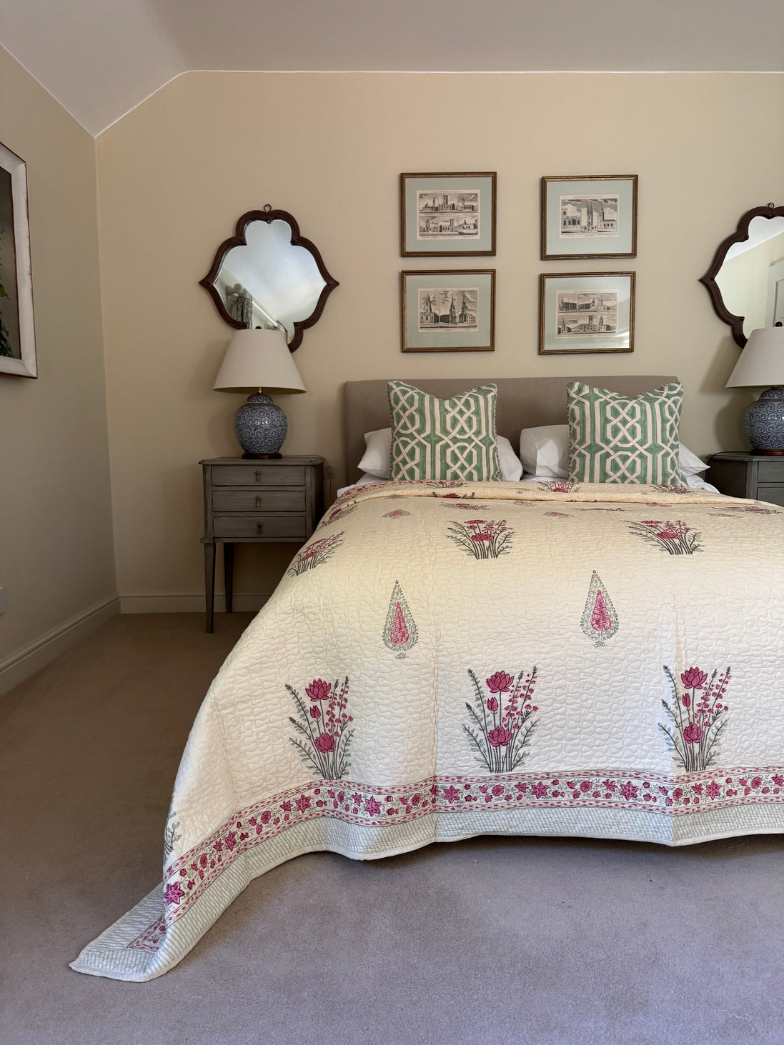 A styled bedroom scene featuring a cream quilted bedspread with pink floral motifs and green geometric accent patterns, styled on a bed with matching pillows in a neutral-toned room.