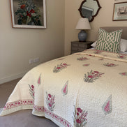 A cream-colored quilted bedspread with pink and green floral patterns is styled on a bed in a bedroom setting, accompanied by a green and white patterned pillow, a bedside table, and wall art.