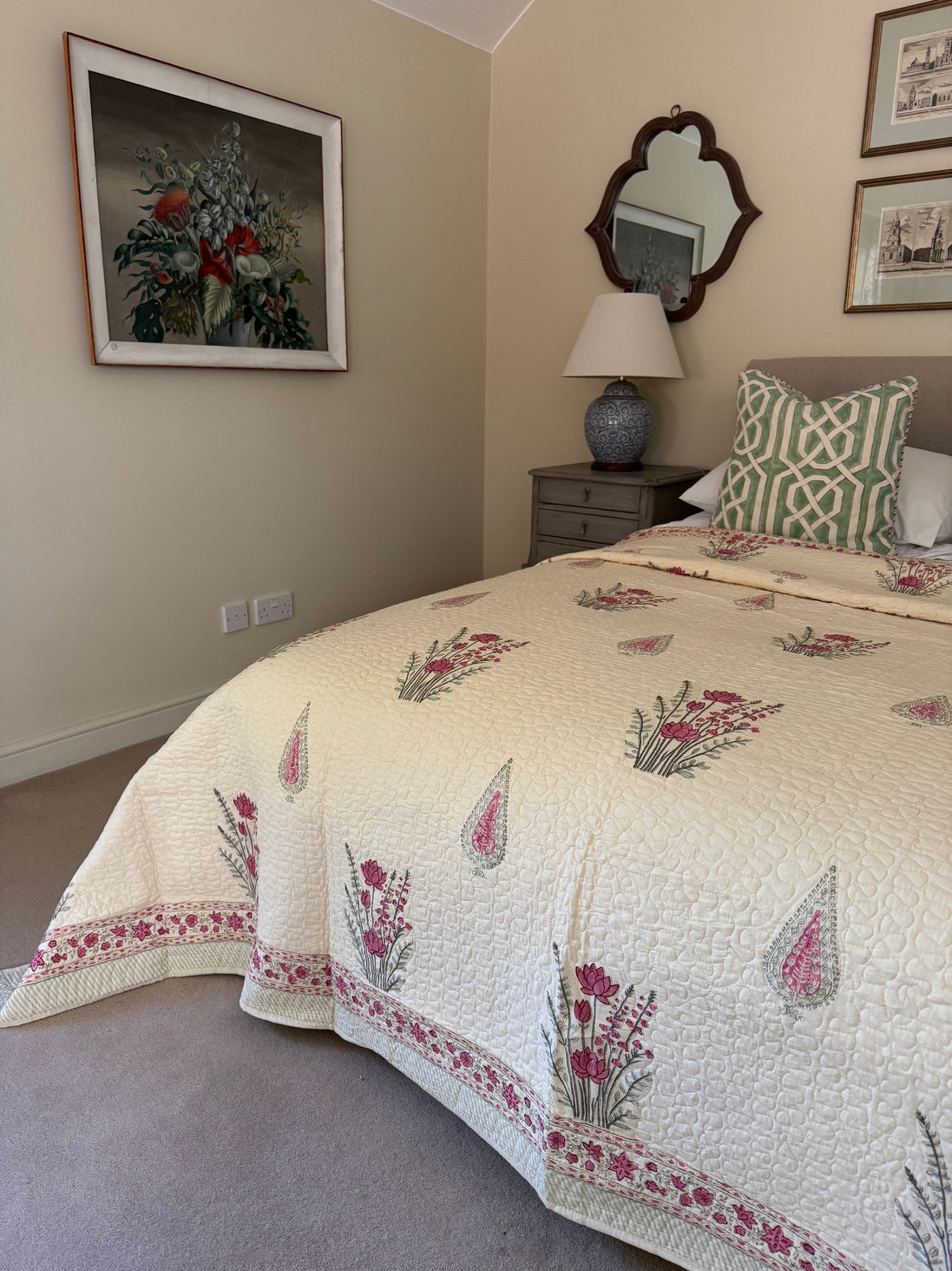 A cream-colored quilted bedspread with pink and green floral patterns is styled on a bed in a bedroom setting, accompanied by a green and white patterned pillow, a bedside table, and wall art.