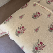 A cream-colored quilted bedspread with a repeating pattern of pink flowers and green leaves, styled on a bed in a bedroom setting.