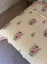 A cream-colored quilted bedspread with a repeating pattern of pink flowers and green leaves, styled on a bed in a bedroom setting.