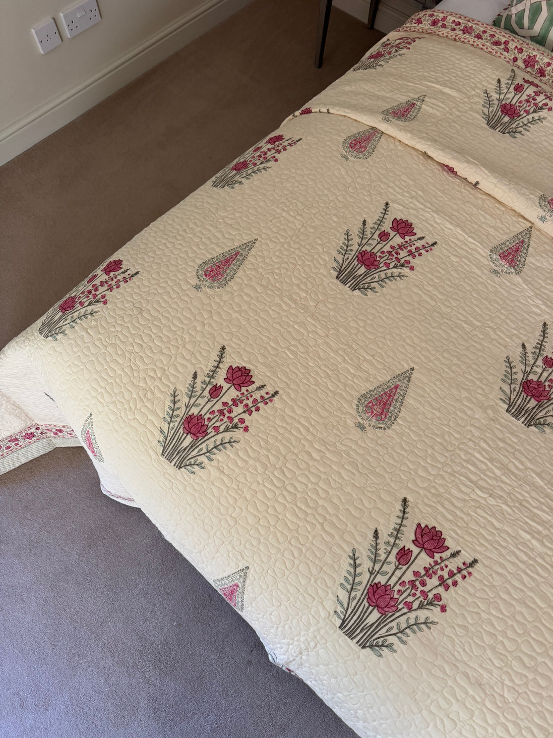 A cream-colored quilted bedspread with a repeating pattern of pink flowers and green leaves, styled on a bed in a bedroom setting.