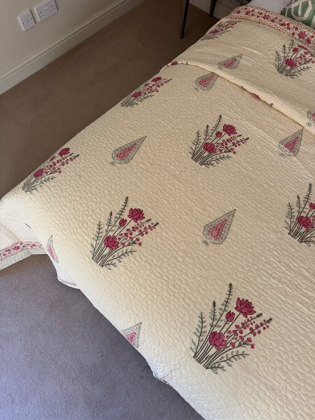 A cream-colored quilted bedspread with a repeating pattern of pink flowers and green leaves, styled on a bed in a bedroom setting.