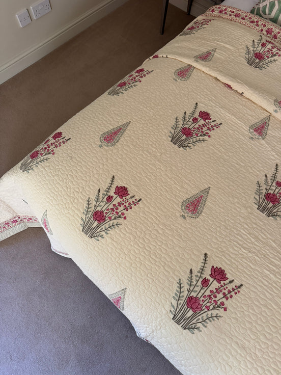 A cream-colored quilted bedspread with a repeating pattern of pink flowers and green leaves, styled on a bed in a bedroom setting.