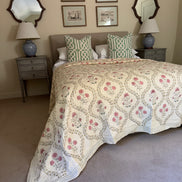 A bed dressed in a cream quilted coverlet with pink floral motifs and green-and-white patterned pillows, styled in a bedroom with matching nightstands, lamps, framed wall art, and mirrors.