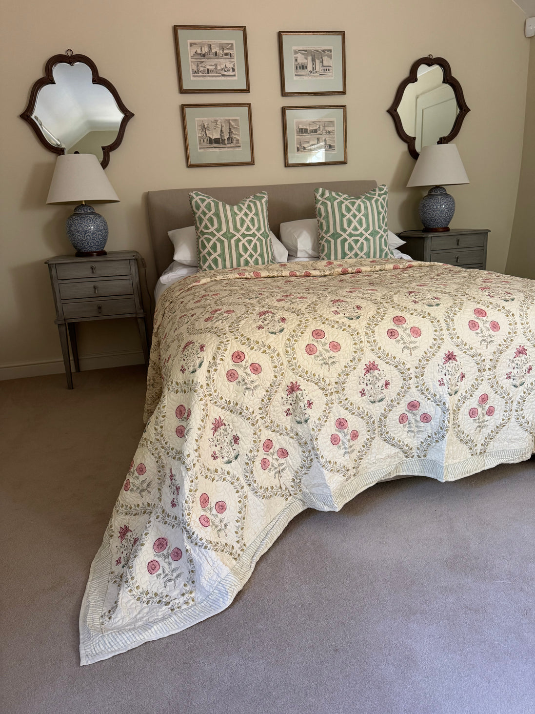 A bed dressed in a cream quilted coverlet with pink floral motifs and green-and-white patterned pillows, styled in a bedroom with matching nightstands, lamps, framed wall art, and mirrors.