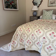 A cream-colored quilted bedspread with a pink and green floral pattern, styled on a bed in a bedroom setting with a matching green and white patterned pillow, a bedside table, and a mirror.