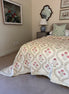 A cream-colored quilted bedspread with a pink and green floral pattern, styled on a bed in a bedroom setting with a matching green and white patterned pillow, a bedside table, and a mirror.