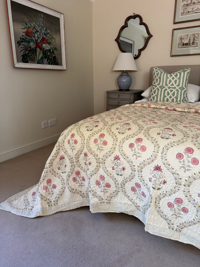 A cream-colored quilted bedspread with a pink and green floral pattern, styled on a bed in a bedroom setting with a matching green and white patterned pillow, a bedside table, and a mirror.