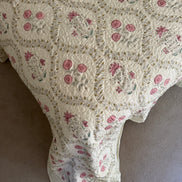 A vintage-style quilted blanket with a cream background, pink floral motifs, and green leaf accents, draped over a bed or chair on a neutral carpet.