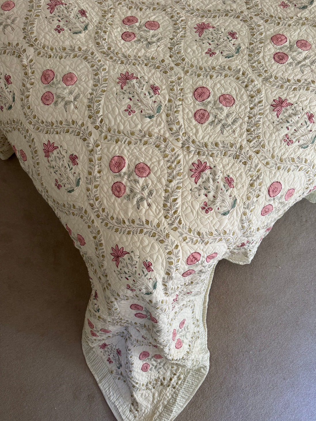 A vintage-style quilted blanket with a cream background, pink floral motifs, and green leaf accents, draped over a bed or chair on a neutral carpet.