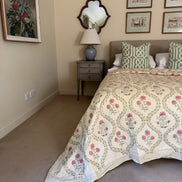 A vintage-style quilted bedspread with a pink and green floral medallion pattern, styled on a bed in a bedroom with a gray nightstand, blue ceramic lamp, and framed wall art.