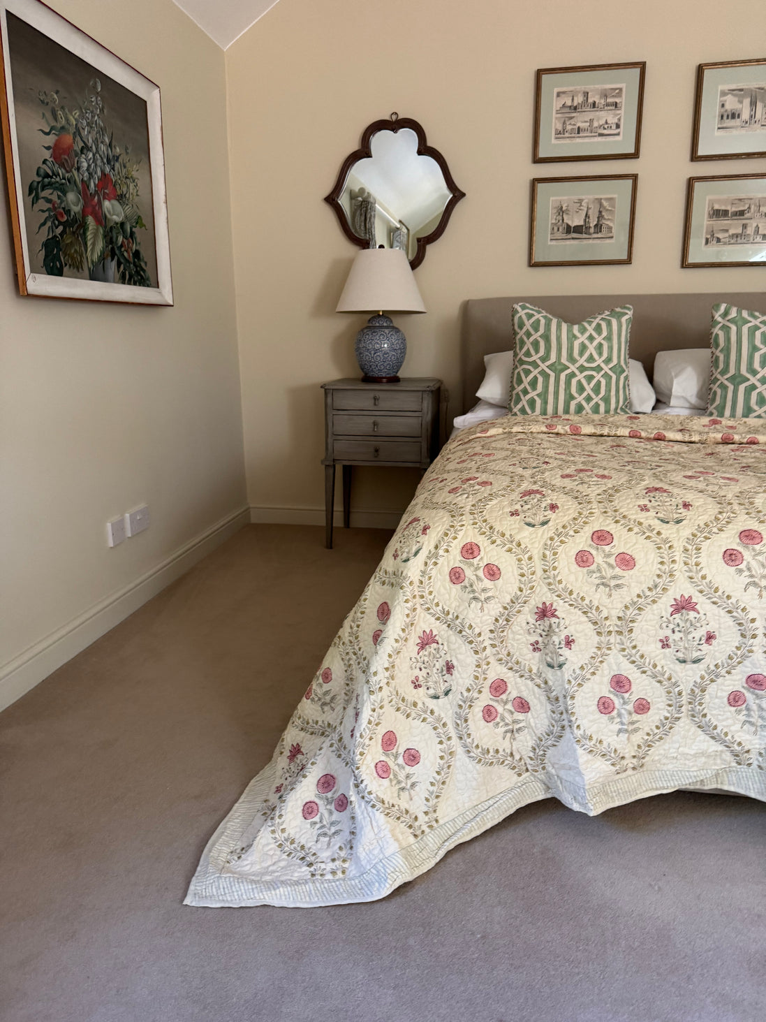 A vintage-style quilted bedspread with a pink and green floral medallion pattern, styled on a bed in a bedroom with a gray nightstand, blue ceramic lamp, and framed wall art.