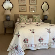 A cream-colored quilted bedspread with red and green floral embroidery and a patterned border, styled on a bed with matching green and white patterned pillows in a bedroom setting.
