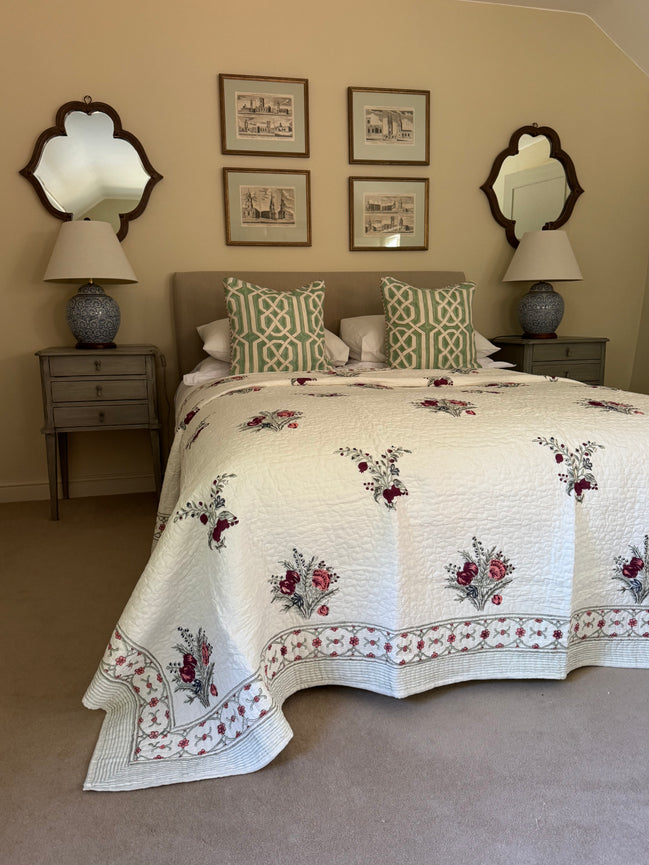 A cream-colored quilted bedspread with red and green floral embroidery and a patterned border, styled on a bed with matching green and white patterned pillows in a bedroom setting.