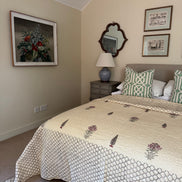 A styled bedroom featuring a bed with a cream and green geometric patterned quilt, matching pillows, a gray headboard, a bedside table with a lamp, and framed wall art on beige walls.
