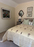 A styled bedroom featuring a bed with a cream and green geometric patterned quilt, matching pillows, a gray headboard, a bedside table with a lamp, and framed wall art on beige walls.