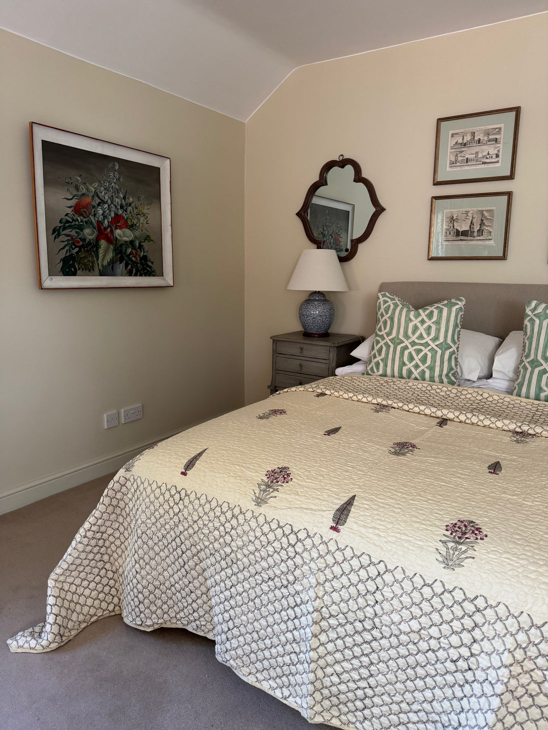A styled bedroom featuring a bed with a cream and green geometric patterned quilt, matching pillows, a gray headboard, a bedside table with a lamp, and framed wall art on beige walls.