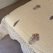 A cream-colored quilted bedspread with embroidered feather and floral motifs, shown on a bed in a bedroom setting.