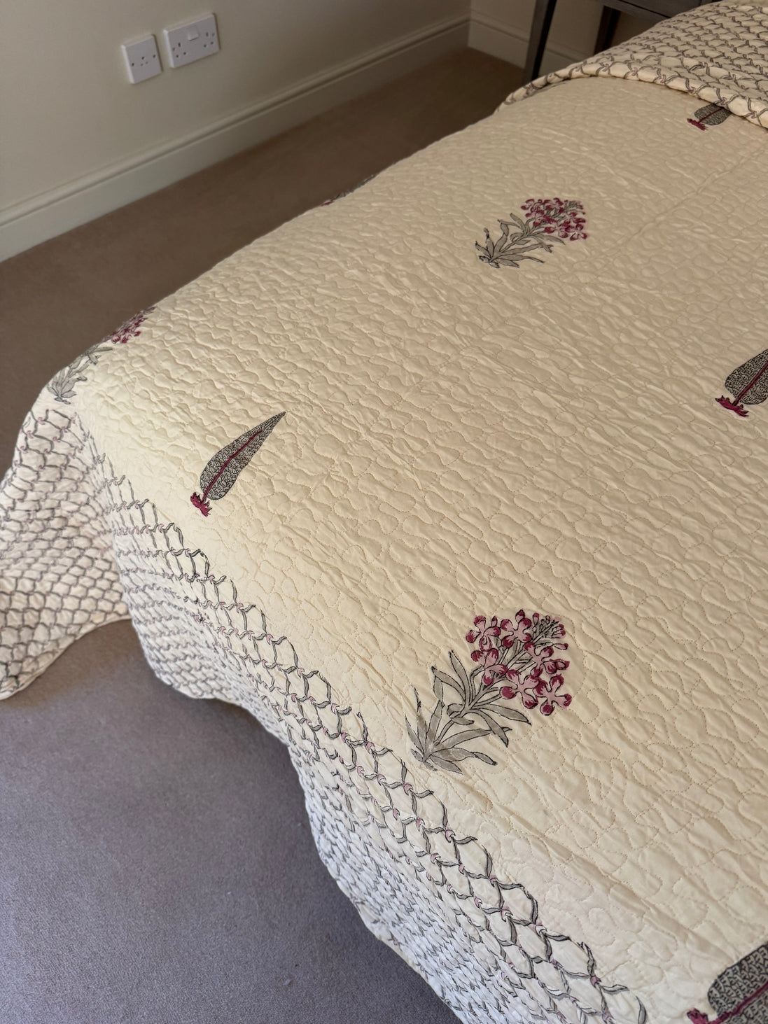 A cream-colored quilted bedspread with embroidered feather and floral motifs, shown on a bed in a bedroom setting.