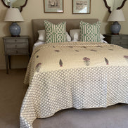 A styled bedroom scene featuring a bed dressed in a cream quilted comforter with a geometric pattern and botanical embroidery, flanked by two green and white patterned pillows. The room is decorated with gray nightstands, blue ceramic lamps, and framed wall art.
