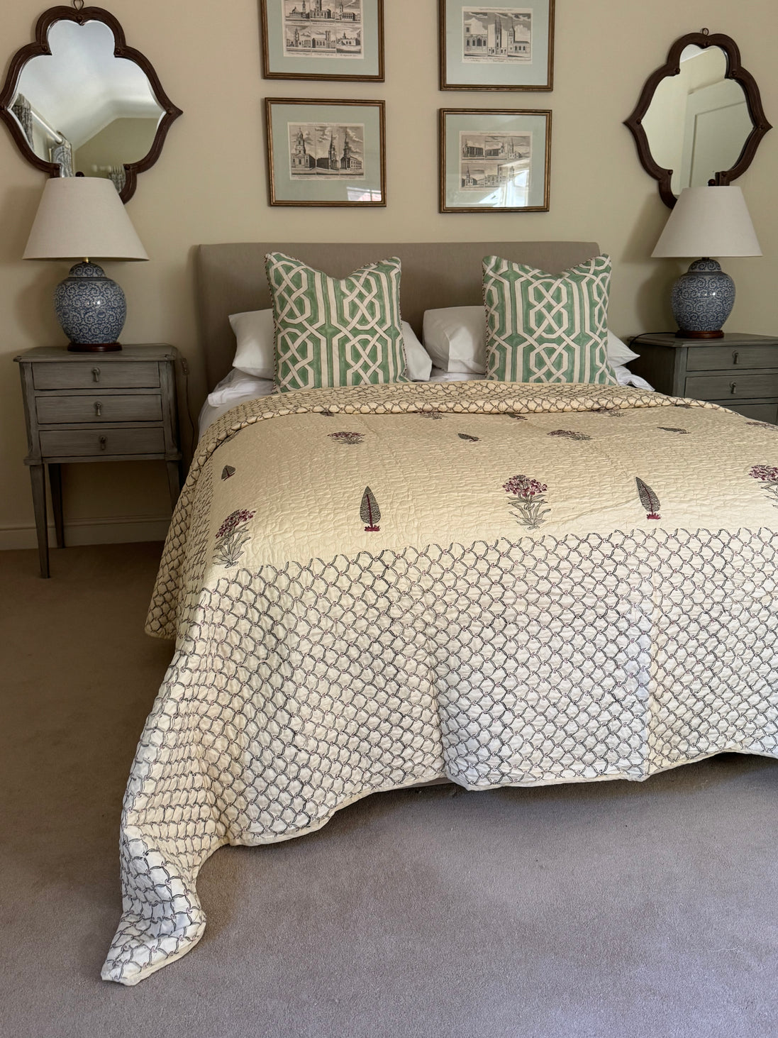 A styled bedroom scene featuring a bed dressed in a cream quilted comforter with a geometric pattern and botanical embroidery, flanked by two green and white patterned pillows. The room is decorated with gray nightstands, blue ceramic lamps, and framed wall art.