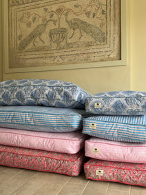 Stack of patterned cushions on a wooden floor with a framed mosaic artwork in the background.